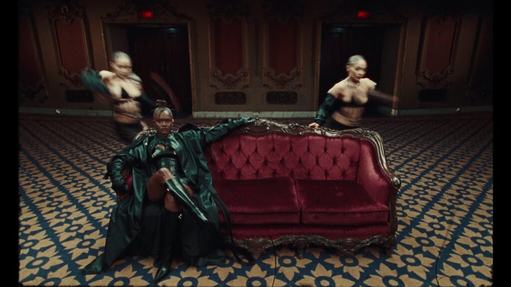 A person in a green coat sitting confidently on a red sofa with two blurred figures dancing in the background. thumbnail for the music video "Amaarae - Angels in Tibet (Official Music Video)"