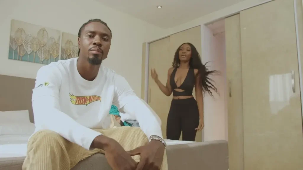 A man sitting on the edge of a bed looking at the camera while a woman enters the room in the background. Thumbnail for "Beeztrap KOTM - Fly Girl feat. Oseikrom Sikanii (Official Video)"