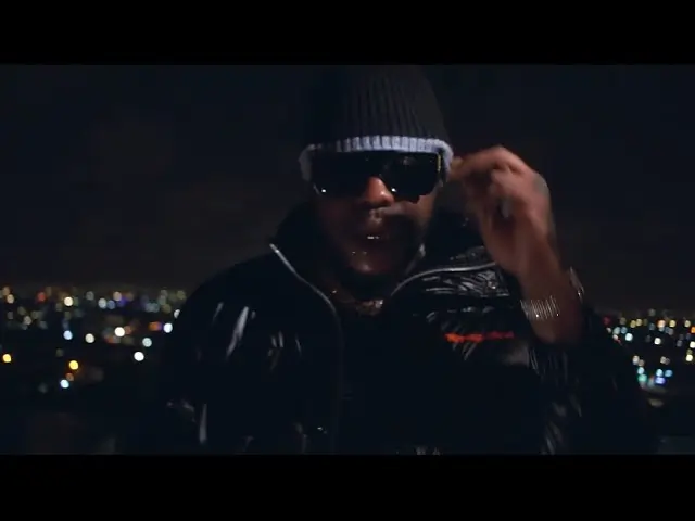 A man in sunglasses and a beanie stands at night with a cityscape behind him. Thumbnail for "Medikal & Uncle Rich - Alpha Hour (Official Video)"