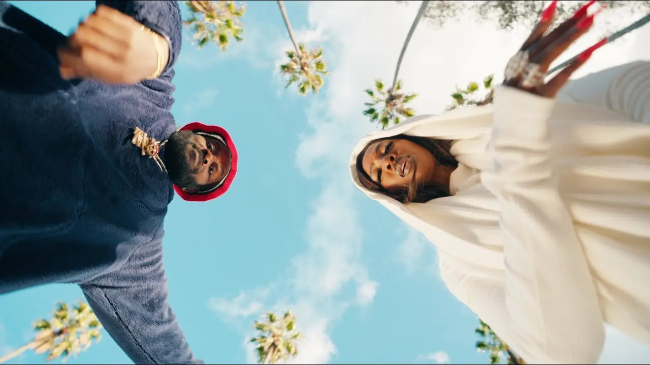 Two individuals dressed in superhero costumes standing back-to-back under a sunny sky with palm trees in the background. Thumbnail for the music video "ODUMODUBLVCK feat. Tiwa Savage - 100 Million (Music Video)"