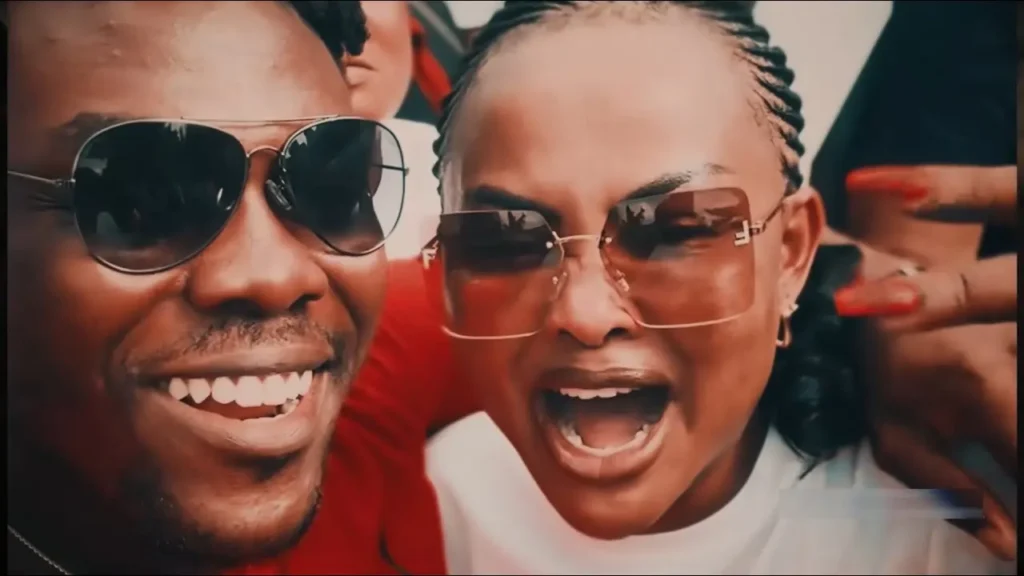 Thubmail for 'Bogo Blay - 24 [VIDEO]'. The image shows Two people (Rapper, Bogo Blay and TV personality, Nana Ama McBrown) wearing sunglasses are smiling and appearing excited in a close-up photo. The person on the right (Nana Ama McBrown) is gesturing with their left hand.