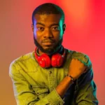 DJ Sleek wearing a green denim shirt and red headphones around his neck stands against a colorful gradient background. Artwork for Article "DJ Sleek nominated for “Best DJ Act Ghana” at the Ghana Entertainment Awards USA 2024"