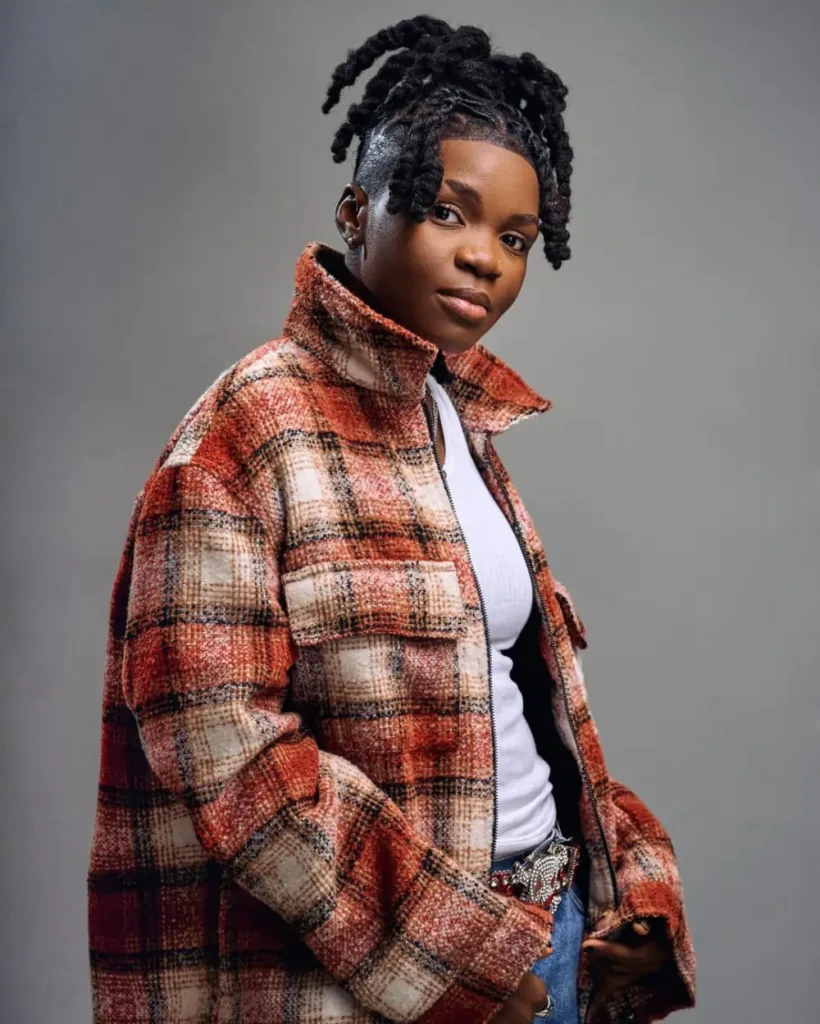 Ghanaian dance, Endurance Grand, with braided hair wearing a red and brown plaid jacket over a white shirt, standing with a neutral expression against a grey background.
