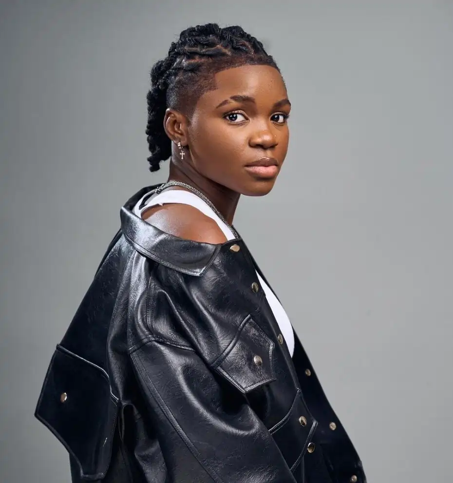 Image of Ghanaian Dancer, Endurance Gold, with braids, wearing a black leather jacket over a white top, looks towards the camera against a gray background.