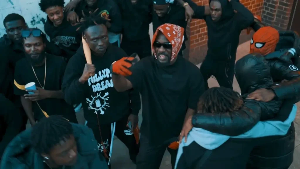 A group of people dressed mostly in black gather outside near a brick wall. One person in sunglasses and a red bandana gestures, while another holds a wooden bat. Some are looking in various directions. . Thumbnail for Jay Bahd - Hate feat. Sarkodie (Official Video)