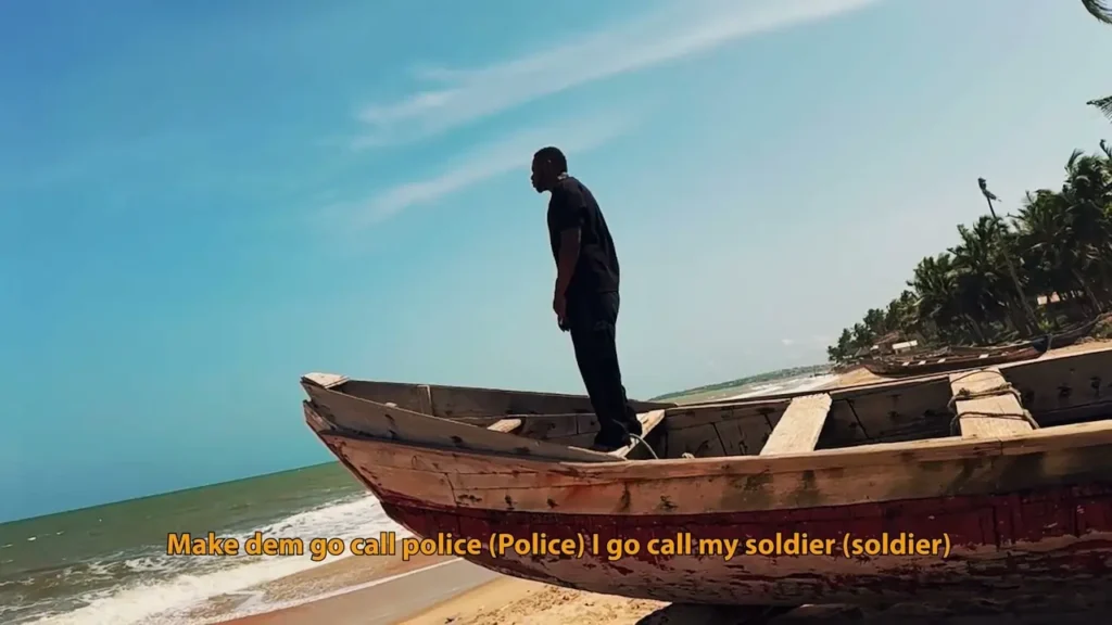 A person in dark clothing stands on a wooden boat on a sandy beach, with the ocean and palm trees in the background. The sky is clear and blue. Medikal - My Story (Visualiser) (Visualiser)