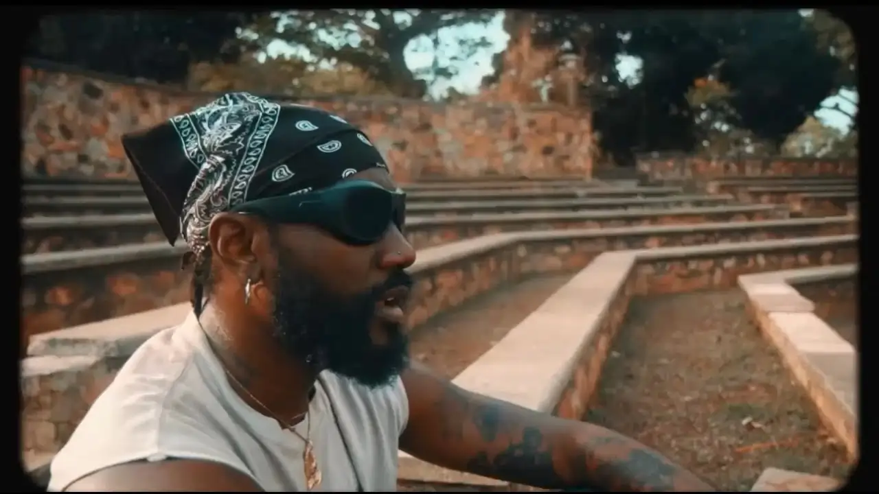 A man with tattoos and a bandana sits on outdoor steps, wearing sunglasses and a sleeveless shirt, in a thoughtful pose. Thumbnail for "Pappy Kojo - Tell 'Em To Shut Up [Feat. Reggie & Skyface Sdw] (Official Music Video)"