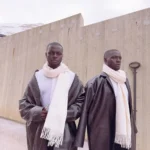 The Mitch Brothers stand in front of a concrete wall wearing white scarves and dark robes, with a snowy mountain backdrop.