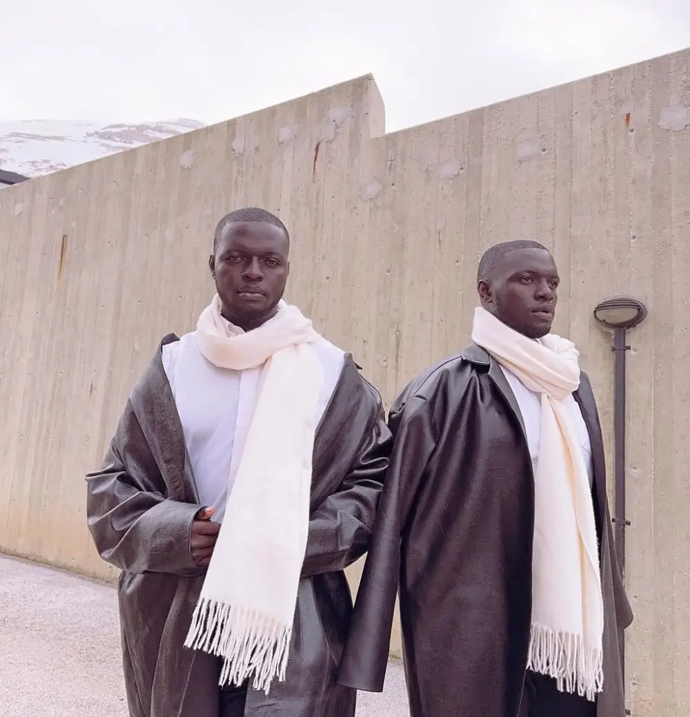 The Mitch Brothers stand in front of a concrete wall wearing white scarves and dark robes, with a snowy mountain backdrop.