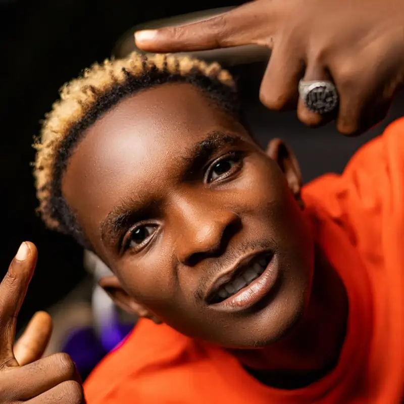 A person with short blonde hair and an orange shirt is pointing both index fingers upwards, with a serious expression on their face. . Baba Tundey officially signs to RuffTown Records