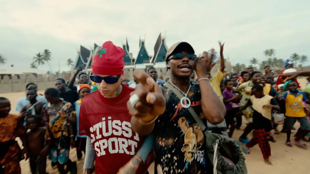 Two people wearing colorful clothes and sunglasses stand in the foreground, surrounded by a lively crowd holding their hands up, reminiscent of scenes from Central Cee - Wave (Official Video), with a distinctive building in the background.