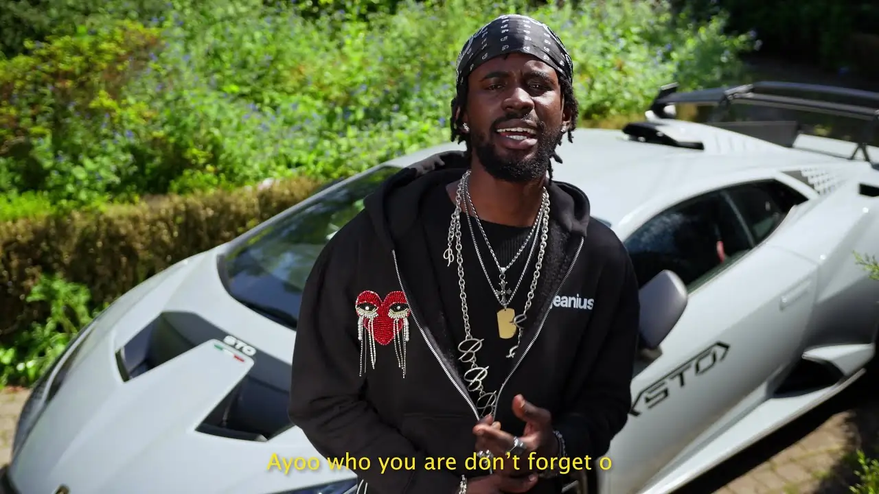 A person with a beard stands in front of a white sports car, speaking. The text on the image reads, "Ayoo who you are don't forget o." Black Sherif - Kilos Milos (Official Visualizer) plays softly in the background.