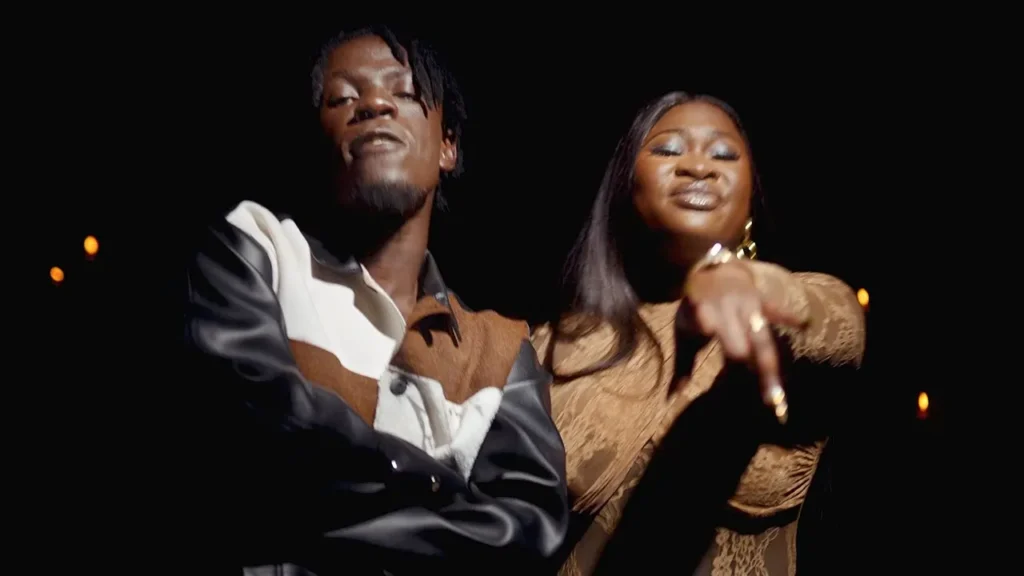 Two people standing close together in a dimly-lit environment, reminiscent of scenes from the "King Paluta - More Money (ft. Sista Afia) (Official Video)." The person on the left has arms crossed, while the person on the right is pointing forward. Both are looking at the camera.