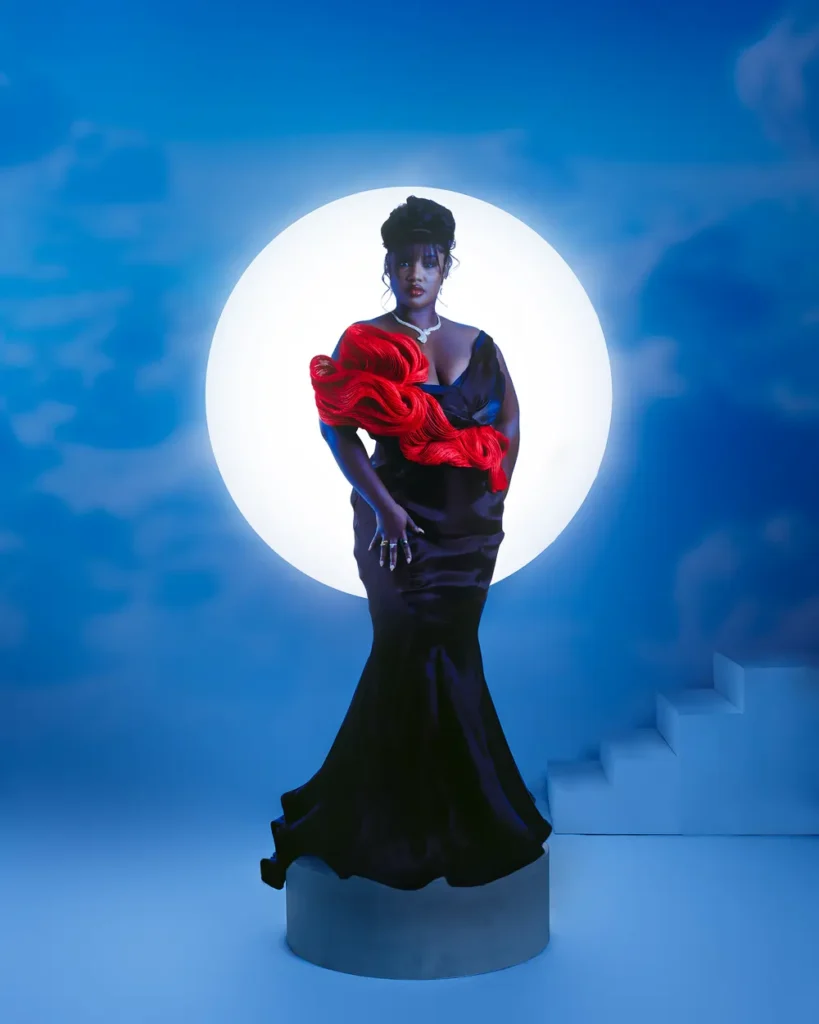Titi Owusu stands on a circular podium at the TGMAs, wearing a black gown with a vibrant red accent. A large white circle is illuminated behind them against a blue sky backdrop.