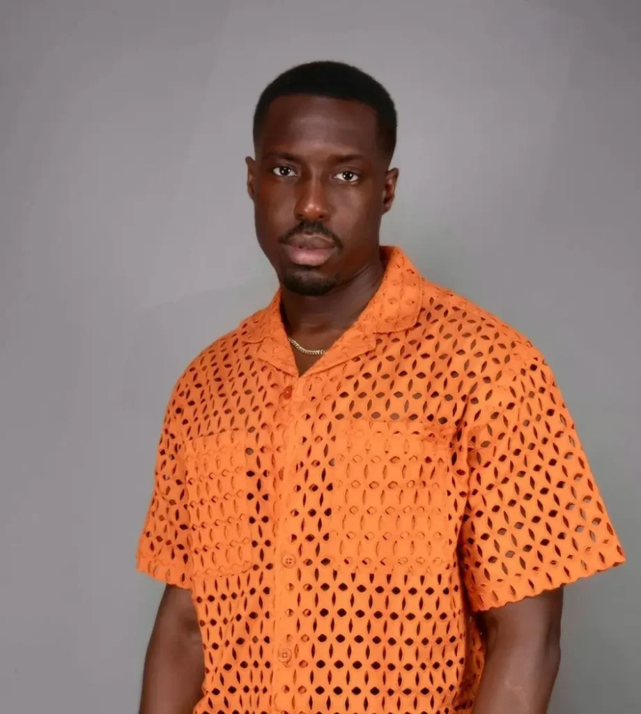 A man wearing an orange, short-sleeved, perforated shirt stands against a plain backdrop. British-Ghanaian star Mark Asari scores big with ‘Energy’ in new Netflix movie “Beverly Hills Cop: Axel F.”