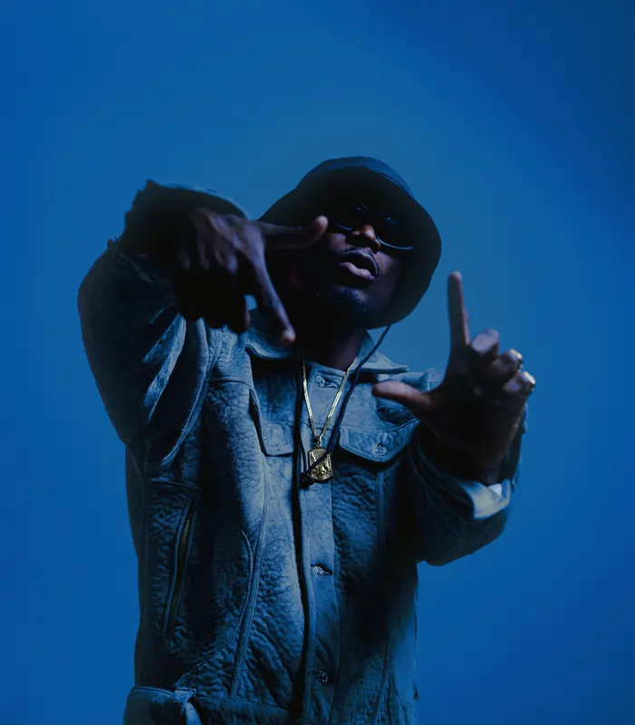 A picture of E.L infront of a blue background. He is in a denim shirt with a bucket hat throwing a sign. Part of promo pics for Bar 7