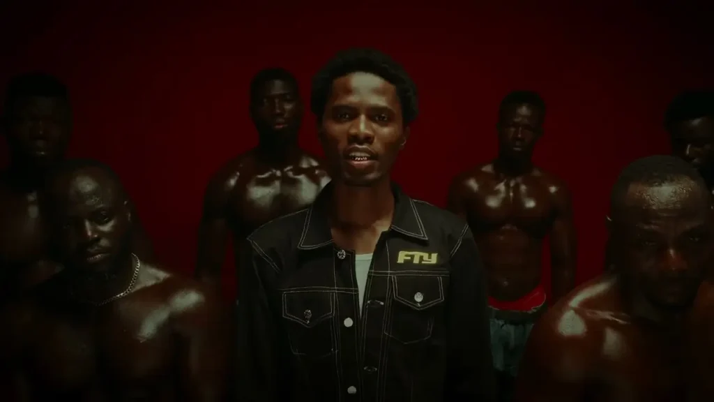 A group of six men are posing against a red background in what appears to be a scene from Kwesi Arthur - Fefe Ne Fe (Official Video). The central figure is wearing a denim jacket, while the others are shirtless, revealing muscular physiques.