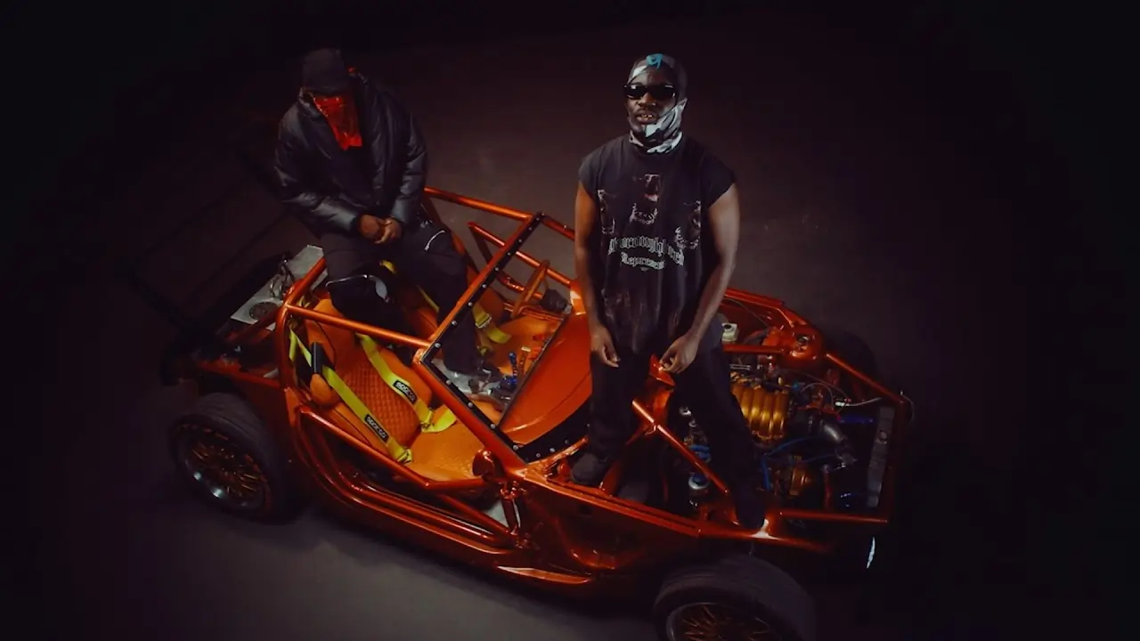 Two individuals stand by a custom-made, open-framed vehicle with a bright orange interior, reminiscent of the bold visuals in Sarkodie - X feat Joey B (Official Video). One person is seated inside the car, while the other stands next to it. The background is dark.