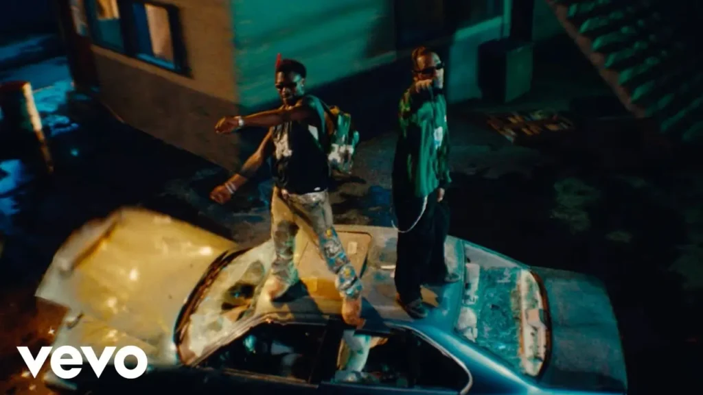 Two individuals stand on the roof of a damaged car in a nighttime setting outside a building. One points forward while the other gestures to the side. The VEVO logo is in the bottom-left corner, hinting at Asake and Travis Scott - Active (Official Video).