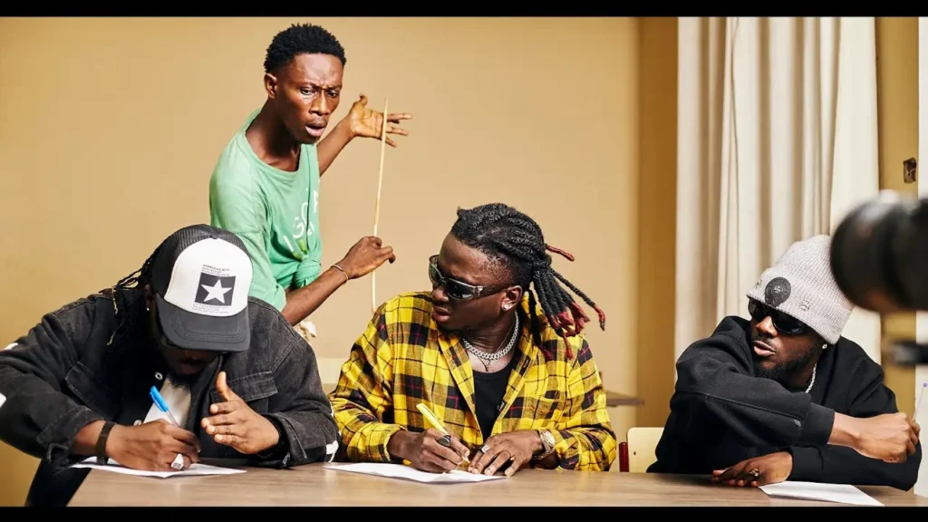 A person in a green shirt points a stick while three individuals wearing hats and dark clothing sit at a table writing, almost like they're working on the next DopeNation - Gbohe (Visualizer).