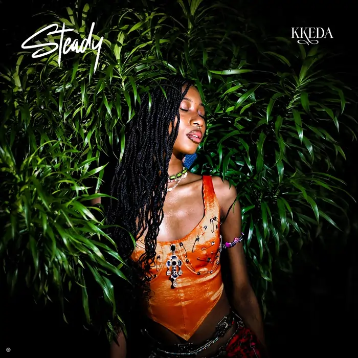 Artwork for post 'Kkeda - Steady'. A woman with long braided hair wears an orange top, standing amidst green foliage. The word "Steady" appears in white at the top left corner, and "KKEDA" stands prominently in white at the top right corner.